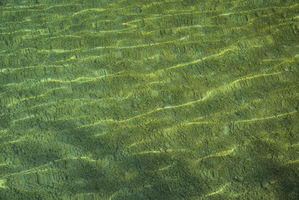Das Foto zeigt eine Kiesfläche am Grund eines sehr klaren Sees, die durch die Wasseroberfläche hindurch sichtbar ist. An der Wasseroberfläche selbst sind keine klassischen Wellen erkennbar, doch die Lichtbrechung erzeugt ein Muster aus sanft gewellten Lichtlinien und Verzerrungen am kiesigen Untergrund. Dadurch wird deutlich, dass sich auf dem ansonsten durchsichtigen Wasser feine Wellen bewegen, die das einfallende Sonnenlicht brechen und so die Struktur des Untergrunds optisch in dynamische, fließende Formen verwandeln.