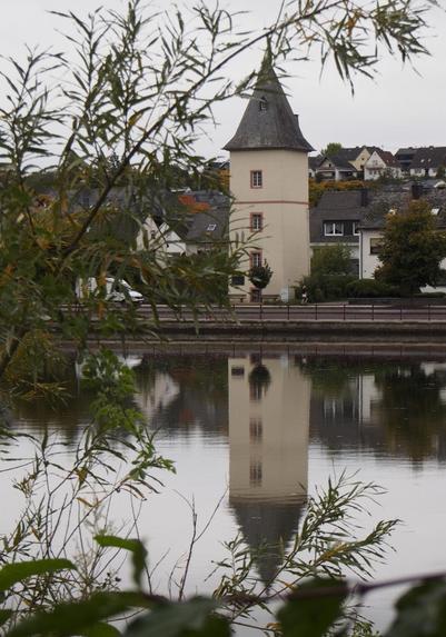 Zwischen den Ästen von Sträuchern sieht man einen cremefarbenen Turm mit spitzem Dach. Auf 4 Etagen ist jeweils ein Sprossenfenster. Der Turm spiegelt sich in der Mosel.