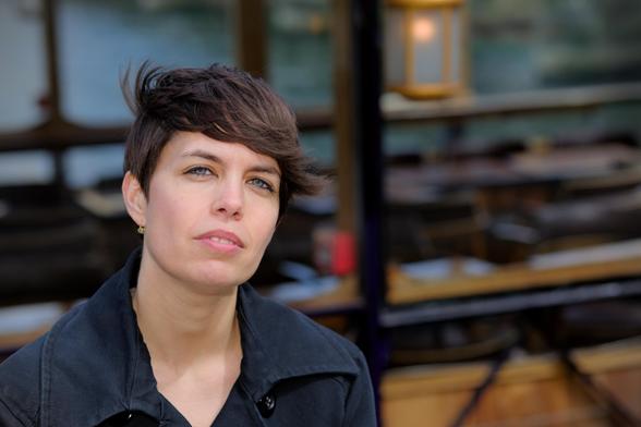a young woman with short black hair, blown by some wind, wearing a black jacket looking badassly at the camera. The background is a piece of a boat with wood and black frames. A warm lamp is in the boat behind a window