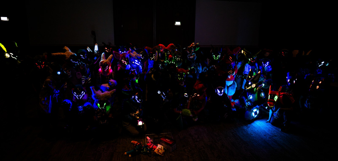 A group of protogen and synth fursuiters sitting in the dark, illuminating only from their LEDs
