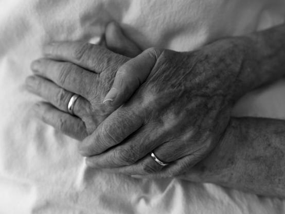 Digital black and white picture of 2 hands of older people. The hands are on top of each other. On both hands wedding rings can be seen. The photo has a shallow depth of field.