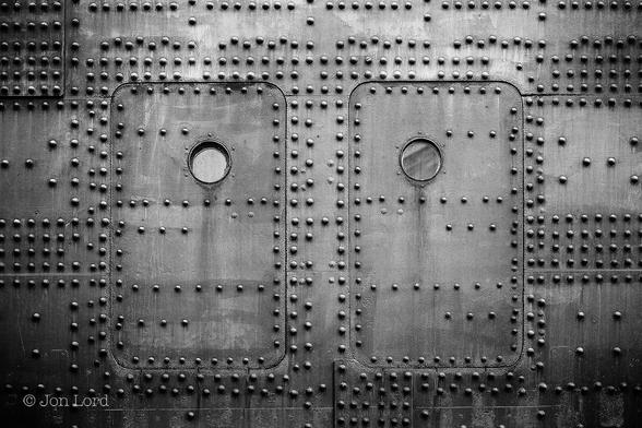 This is an abstract black and white photo in landscape format of the side of the RMS Queen Mary with two small hatches, two small portholes and a multitude of rivets. Long Beach, California (2015).
The entire image is filled with the ink black steel hull of the side of a ship. To the left and right of centre are the two small rectangular hatches set into the side of this large passenger ship. Each hatch is about 1.8m high by a little more than a metre wide. About three quarters of the way up, in each hatch, is a small porthole offset slightly from centre. All around the image are hundreds of large round head rivets, many in neat rows and lines, others randomly located and spaced. The hull, hatches and rivets belong to the RMS Queen Mary, Launched in Glasgow, Scotland In 1934 and has been moored in Long Beach, California since 1967 as a hotel, museum and film location.
