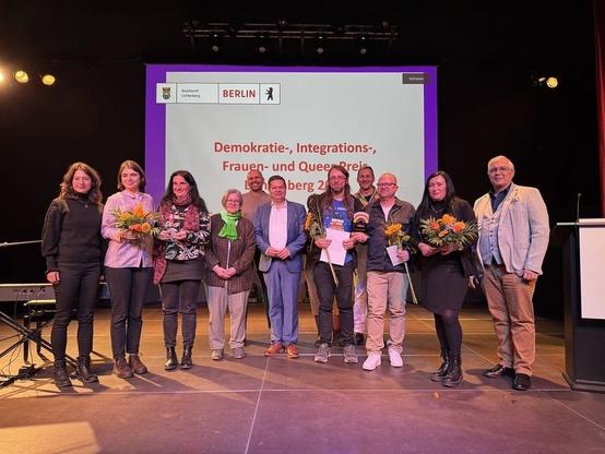 Auf dem Foto steht eine Gruppe von elf Personen auf einer Bühne vor einer großen Leinwand. Die Leinwand im Hintergrund zeigt ein lila-weißes Design mit dem Berliner Wappen und dem Text "Demokratie-, Integrations-, Frauen- und Queer Preis" und der Angabe des Bezirks Lichtenberg.

Mehrere Personen halten kleine Blumensträuße in Gelb- und Orangetönen. In der Mitte der Gruppe steht ein Mann in einem hellblauen Anzug, der Bezirksbürgermeister.