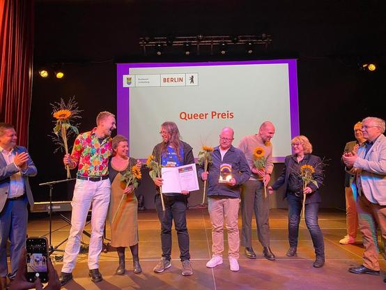 Dieses Bild zeigt eine Gruppe von neun Personen auf einer dunklen Bühne bei einer Preisverleihung. Im Hintergrund ist eine große, beleuchtete Leinwand zu sehen, die das Wappen von Berlin und Lichtenberg sowie den Text "Queer Preis" in Rot zeigt.

Im Zentrum der Gruppe stehen die Preisträger, die sichtlich fröhlich sind. Ein Mann in einem auffälligen, bunten Hemd hält einen großen Strauß mit Sonnenblumen und freut sich zusammen mit einer Frau an seiner Seite. Zwei weitere Preisträger halten ihre Auszeichnungen: ein Mann mit langen Haaren hält eine Urkunde und ein anderer Mann mit Brille hält einen glänzenden, regenbogenfarbenen Pokal. Mehrere Personen halten Sonnenblumensträuße. Am linken und rechten Rand des Bildes stehen weitere Personen, die applaudieren oder zusehen. Im Vordergrund rechts unten ist eine Hand zu sehen, die ein Smartphone hält und damit ein Foto oder Video aufnimmt.