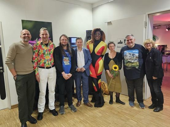 Mitglieder der AG Queer zusammen mit der Queerbeauftragten und dem Bezirksbürgermeister von Lichtenberg. 

Das Bild zeigt eine Gruppe von acht Personen, die eng beieinander in einem Innenraum mit hellem Holzboden und weißen Wänden stehen und in die Kamera lächeln. Die Gruppe ist vielfältig, besonders hervorstechend ist eine Person in der Mitte in einem auffälligen, enganliegenden, gelb-rot-schwarzen Anzug. Zwei Männer tragen ebenfalls farbenfrohe Kleidung, darunter ein Mann mit einem bunten, gemusterten Hemd. Eine Frau hält eine Sonnenblume.