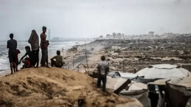 A wide shot shows a group of people standing on a sandy hill overlooking a beach. The beach is covered in debris and wreckage, stretching into the distance towards a cityscape on the horizon. Several people are gathered on the hill, looking out towards the ocean and the wreckage, with one person kneeling. In the foreground, there are dark, rectangular objects, possibly tarps or debris, arranged in a pile. The sky is overcast and muted, contributing to a somber atmosphere.