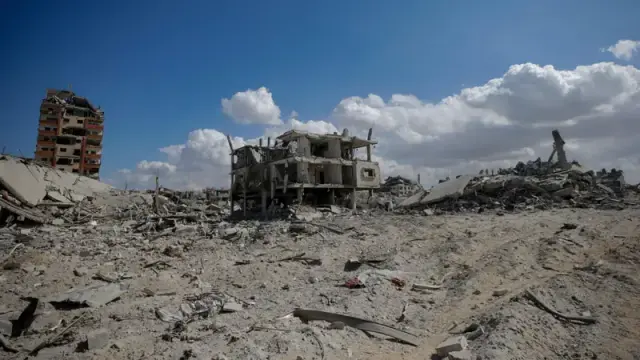 A wide shot depicts a landscape covered in rubble and debris, likely the aftermath of a destructive event. The foreground and middle ground are filled with a vast expanse of gray, fragmented materials—concrete, wood, and other building components—scattered across the terrain. A partially standing, multi-story building with a flat roof and open windows is visible in the center, appearing heavily damaged and incomplete. To the left of this structure, a taller, more intact building with orange accents stands in the distance. The sky is a clear blue with scattered white clouds, contrasting with the devastation below.