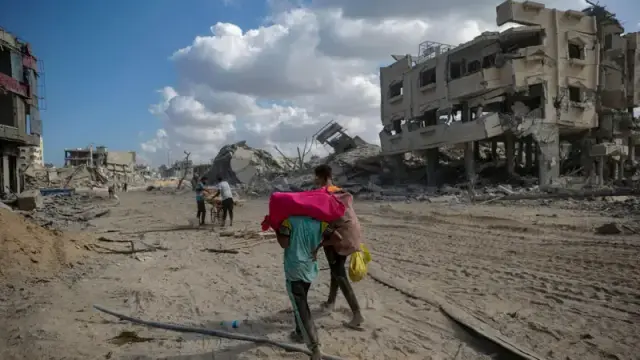 A wide, outdoor shot depicts a scene of widespread destruction, likely from a bombing or similar event. The ground is covered in a layer of debris and dust, with rubble from buildings scattered throughout. In the foreground, a person is carrying a bundle of pink fabric on their back, with a yellow object dangling from one hand, walking away from the camera. Further back, other people can be seen among the ruins, seemingly moving through the area. The buildings in the background are heavily damaged, with many appearing partially collapsed and exposing interior rooms. The sky is bright blue with scattered white clouds.