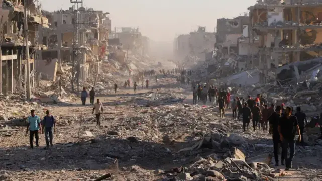 A long, wide street stretches into the distance, flanked by heavily damaged buildings on both sides. The street is covered in rubble, debris, and remnants of structures, creating a desolate and chaotic scene. Numerous people are walking along the street, moving in both directions, appearing small in comparison to the surrounding destruction. Buildings are mostly tan and brown, with visible damage like holes, broken walls, and crumbling facades. The light is somewhat hazy, casting a muted tone over the landscape and creating a sense of distance and bleakness.
