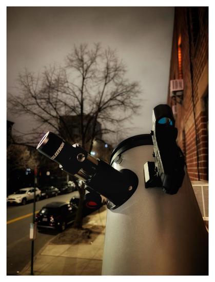 Photo of the gray optical tube of a telescope, with a Celestron eyepiece and attached finder scope, pointing up at a featureless, completely overcast night sky. In the background are a red brick apartment building, a street with parked cars, and a tree with bare branches.