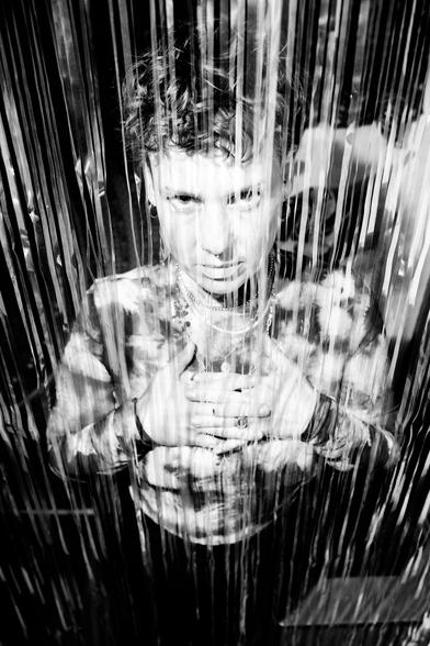 Schwarzweiß Foto von Claudia Döffinger mit verschränkten Händen auf der Brust. Von Oben durch durch hängende Streifen am Türrahmen fotografiert.