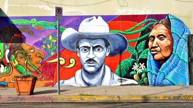 Mural showing Farabundo Marti in a broad brimmed hat, next to a modern indigenous woman, in a blue shawl, and ancient depiction of an indigenous man, with a long-tailed green Quetzal bird in the background.