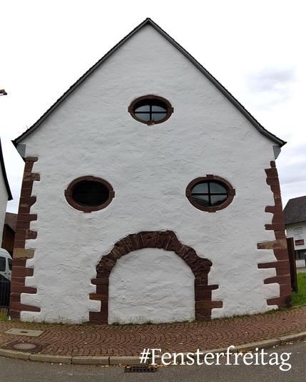 Die weiß verputze Fassade einer Kirche, Stirnseite mit spitzem Giebel, die Kanten links und rechts aus unterschiedlich großen roten Sandsteinen. Drei Fenster oben in einem Dreieck angeordnet, in der Form liegende Ovale. Das Fenster oben und unten rechts haben Scheiben und ein dunkles Fensterkreuz, das Fenster unten links ist offen bzw. der Blick hinein schwarz. Unter den Fenstern am rot gepflasterten Gehweg ist in der Fassade ein steinerner Torbogen, der allerdings zugemauert und ebenfalls weiß verputzt ist. Der Himmel hinter dem Gebäude ist weiß mit einigen blauen Flecken.