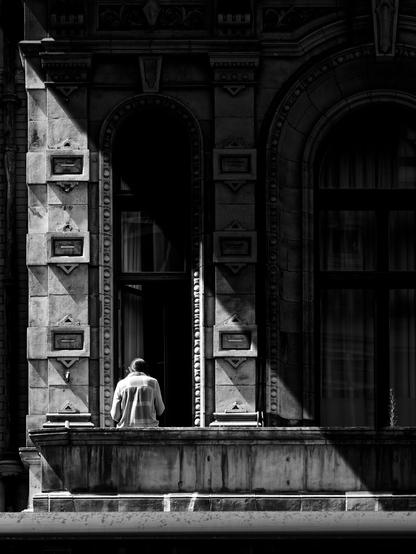 Black and white photo of a person standing on a balcony, framed by the ornate stone architecture of an old building. Sharp sunlight and deep shadows create a dramatic contrast across the facade.