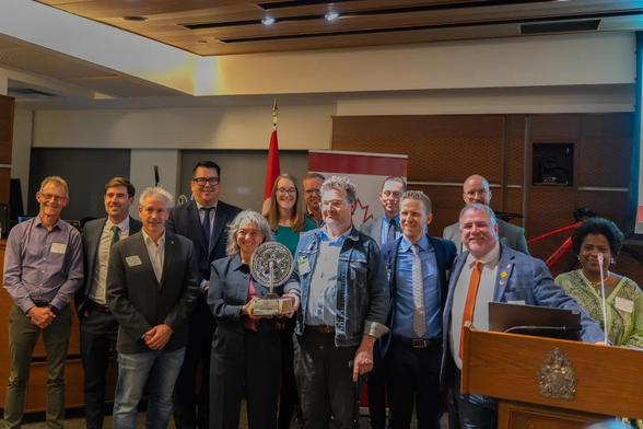 There are thirteen smiling people in a group posing for this photo. In the front, a woman is presenting an award to a man. The award is a wooden base with a bicycle from crank on it.