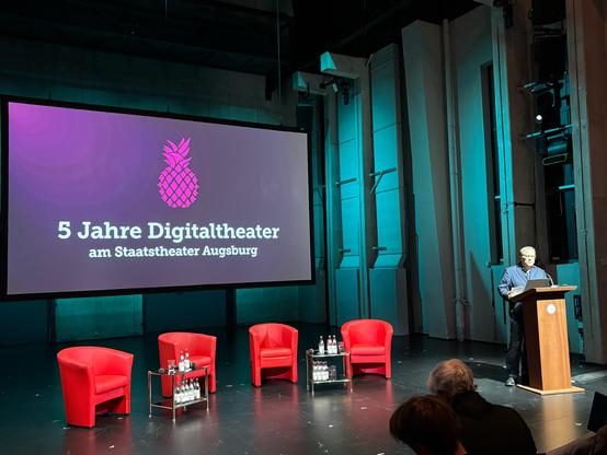 André Bücker - Staatsintendant des Staatstheater Augsburgs: Auf einer Bühne steht ein Mann hinter einem Pult, rechts im Bild. Links dominiert eine große Leinwand mit der Aufschrift: „5 Jahre Digitaltheater am Staatstheater Augsburg“. Darüber ist ein pinkes Symbol einer stilisierten Ananas zu sehen. Drei rote Sessel und kleine Glastische mit Wasserflaschen stehen bereit.