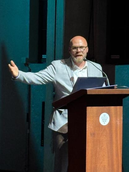 Dr. Fabian Mehring - Digitalminister Bayerns: Ein Mann in einem hellgrauen Anzug steht hinter einem hölzernen Rednerpult auf einer Bühne. Er spricht engagiert, eine Hand ist weit ausgebreitet, die andere ruht auf dem Pult. Das Bühnenlicht taucht ihn in ein türkisfarbenes Licht, der Hintergrund ist dunkel.
