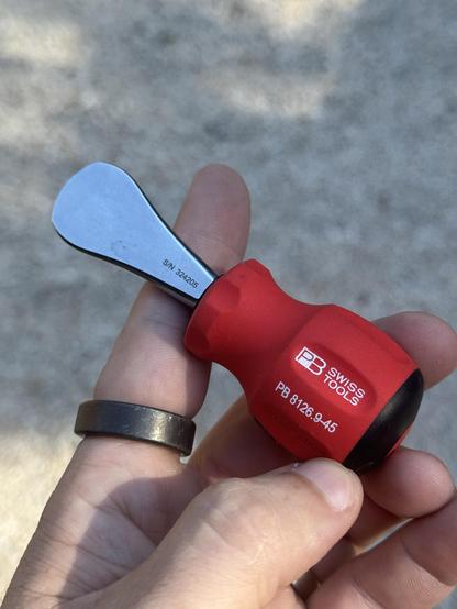Red handled pb Swiss stubby “coin driver” up close. Placed in the left hand with black outs ring on the index finger in front of blurred crushed rock in the background. The blade on the left of the driver is shiney and the tip resembles the shape of a US quarter