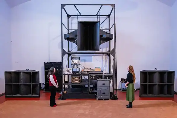 Photo: complex stereo set-up (with both vinyl & reel-to-reel elements) surrounded by over-sized horn speakers (salvaged from cinemas) with a large metal frame suspending a central speaker above the stereo. (Two curators stand next to it for size comparison - the speakers are each about the size of a person)