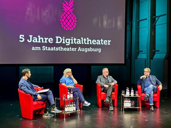 Vier Personen sitzen auf einer Bühne in roten Sesseln und führen ein Gespräch. Hinter ihnen zeigt eine große Leinwand das Logo einer pinken Ananas und den Schriftzug „5 Jahre Digitaltheater am Staatstheater Augsburg“. Zwischen den Sesseln stehen Glastische mit Wasserflaschen und Gläsern. Die Personen wirken konzentriert und engagiert im Austausch, die Bühne ist in türkisfarbenes Licht getaucht. Die Szene vermittelt eine feierliche und zugleich sachliche Atmosphäre.