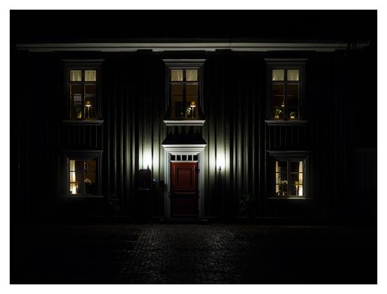 En liggande bild på ett grönt trähus i mörker. Fem fönster är svagt upplysta och två lampor sitter på varsin sida om den bruna trädörren.