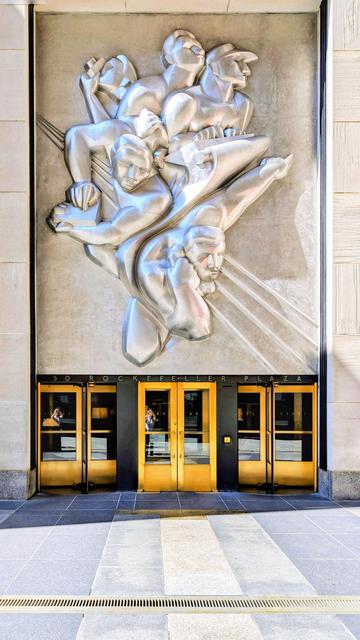 A view of the entrance to 50 Rockefeller Plaza in New York City. A large, stylized silver relief sculpture is mounted above the golden revolving doors.