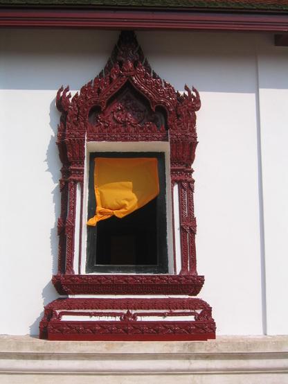 Eine sonnengelbe Gardine etwas zusammengerafft in einem offenen Fenster. Der Fensterrahmen ist aufwändig geschnitzt und rot gestrichen. Er hebt sich von der weißen Wand ab. In diesem Bild herrschen vier Farben vor: gelb, rot, weiß und schwarz.