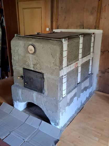 Gray plastered oven sideview with oven sides with iron support frame.
Old oven door fitted in front of the oven entrance. The old lid of the claning opening fitted above the oven door. Terracotta tiles visible in front of the ofen with two wooden templates for the round arch on it. A old wooden paneling is visible on the left, top and right edges of the photo. A wooden parquet floor is visible to the left and right of the stove and a wooden sliding door with a light switch in its frame on the left side of the oven.