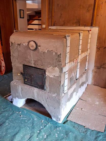 Gray plastered oven sideview with oven sides with iron support frame.
Partly new plaster layer in beige color from above shows ongoing plaster work. A green tile lies on the floor to protect the floor from mortar.
Old oven door fitted in front of the oven entrance. The old lid of the claning opening fitted above the oven door. A wooden sliding door with a light switch in its frame on the left side of the oven.