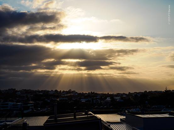 Not long after sunrise the sun's light breaking through ragged clouds producing many shafts of golden light from an intense, diffuse glow. The light picks out the skyline and gleams off the river below. Blue sky is just visible high in the corners of the image.