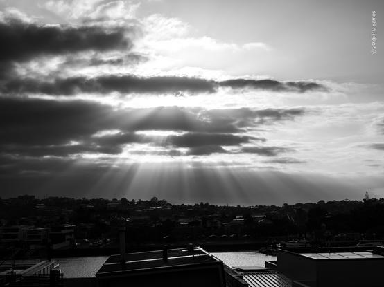 Not long after sunrise the sun's light breaking through ragged clouds producing many shafts of light from an intense, diffuse glow. The light picks out the skyline and gleams off the river below.