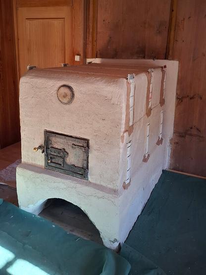 Beige plastered oven sideview with oven sides with iron support frame.
New plaster layer in beige shows ongoing plaster work. A green tile lies on the floor to protect the floor from mortar.
Old oven door fitted in front of the oven entrance. The old lid of the claning opening fitted above the oven door. A wooden sliding door with a light switch in its frame on the left side of the oven.