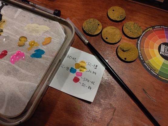 Some colors (yellow, two tones of gold, a teal , a fluorescent pink and a fuchsia, painted on a sheet of paper with paint numbers next to them. 
Around that paper are painting supplies, like a wet palette with the colors, a brush, the finished bases and a color wheel sticker, everything on a wooden surface