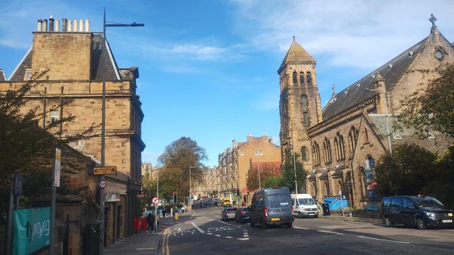 Looking north across Holy Corner on a day of golden, autumn sunlight & blue skies, the church tower of the Eric Liddell Centre and Morningside United Church visible on the eastern side, along with stone, Victorian tenements
