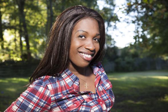A photo shows a Black woman, Ashley Diamond, wearing a plaid shirt and long brown hair. She smiles warmly, looking directly at the camera. In the background is a shaded grove of trees. End image description.