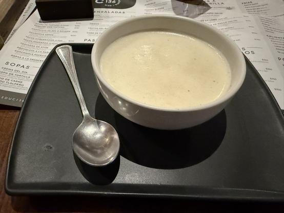 A bowl of creamy soup sits on a black plate, accompanied by a silver spoon. In the background, a partially visible menu can be seen.