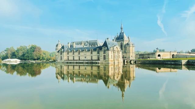 Le chateau de Chantilly qui se reflète dans une étendue d’eau. Le ciel est bleu. L’eau calme.
