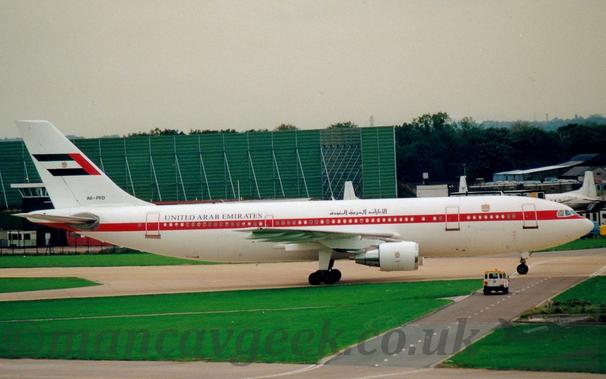 Side view of a twin engined jet airliner taxiing from left to right.
The plane is largely white, with a red stripe running along the body, covering the passenger cabin windows.
There are black "United Arab Emirates" titles on the upper fuselage, in an Arabic script above the wings, and in English at the rear.
The black registration "A6-PFD" is on the base of the tail, which has a large United Arab Emirates flag in the middle.
Lush green grass fills the foreground, with a black roadway splitting it on the grass, with a white vehicle with a yellow roof waiting for the plane to taxi past.
A large green metal frame dominates the background on the left, with the tail of a smaller white airliner nestled amongst small buildings, with trees behind.
Bright but hazy sky fills the rest of the frame.