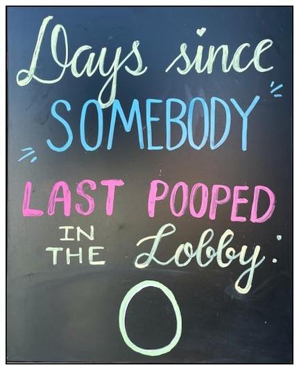 Black chalkboard with white, pink, and blue chalk handwriting displayed. The message humorously counts "0 days since somebody last pooped in the lobby," with each word in a different size and style of writing. A small heart is drawn above the words.