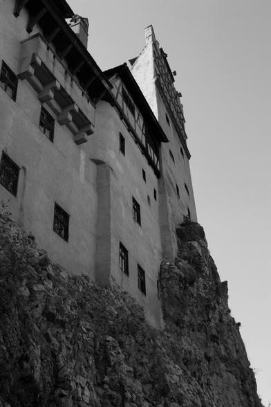 Black and white image looking up at a castle sitting on top of very steep rocks. It's like the castle grew out of the rocks. Dracula's Castle, AKA Bran Castle in Transylvania, Romania.