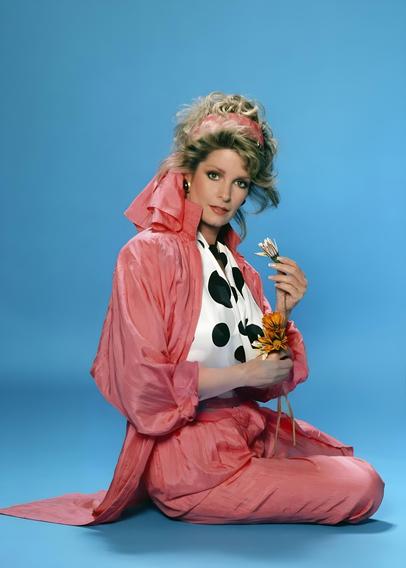 Professionally shot mid-late 1980s portrait of Deidre Hall, known for her iconic role as Marlena Evans on Days of Our Lives. She wears a vibrant pink shell suit over a black-and-white polka dot blouse, with her softly layered, shoulder-length light brown hair tied up in a matching pink headband. Seated against a light blue background, she holds a small bouquet of flowers—including a yellow-orange daisy and white buds—in her hands. Her expression is gentle and composed, gazing directly at the camera. The image evokes nostalgic glamour, soap opera elegance, and the bold, sometimes questionable fashion choices of the era.