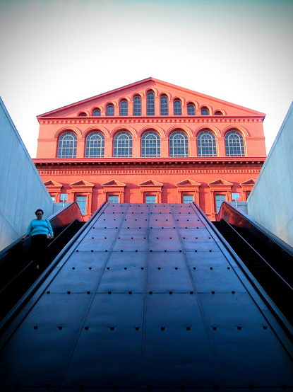 The view of a classical looking building that is painted copper orange, looking upwards through the wide entrance to the dc metro with escalators to each side. The windows are especially beautiful, since the top row with curved tops follow the slope of the roof, the middle row with curved tops are have a grid of leading, and the bottom are adorned with decorative elements around them.