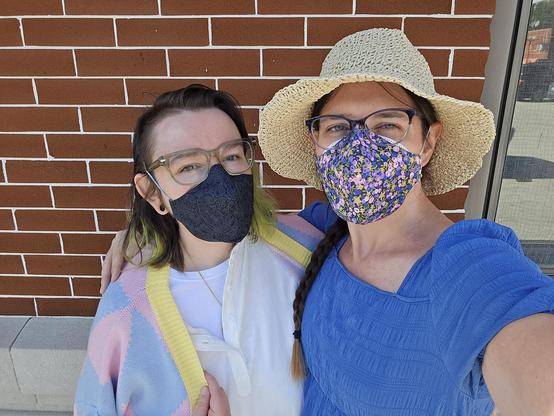 Photo of a white person dressed as Howl Pendragon wearing a black cover over a 3M Aura hugging a white woman dressed as Sophie from Howl's Moving Castle wearing a purple floral mask cover oer her N95 bifold.