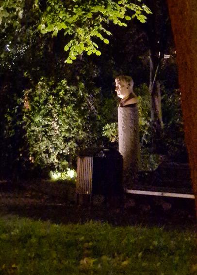 A white stone bust of an older man is lit in a dark park, near bushes and a bin
