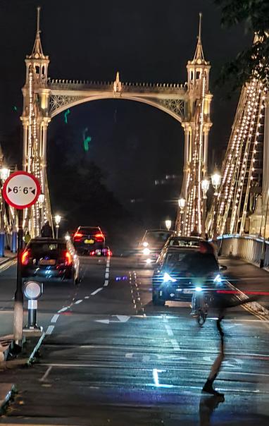 View over Albert Bridge, lit up at night. Crossing the road in front of some cars is what looks like part of an impossibly thin figure - a foot, connecting to a leg and arm - but only a blur where body and head should be.