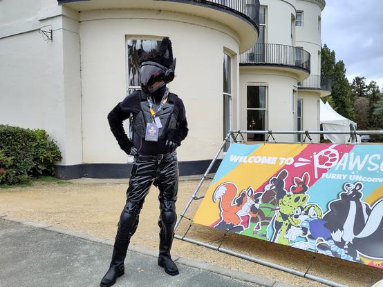 Fursuit photo showing Quirk the protogen standing next to the Pawsome sign outside the hotel.  He is wearing chest and leg armour, PVC jeans and leather boots.
