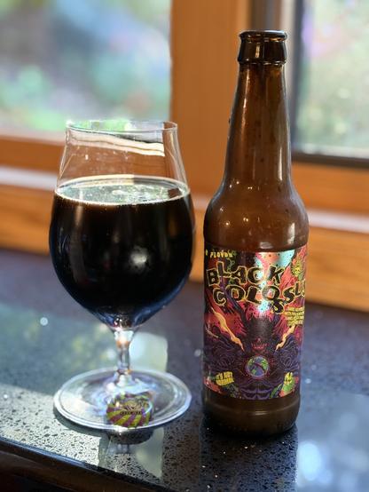 A tall snifter full of dark beer next to a brown bottle with a chromatic colored label and an evil beast
