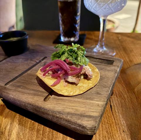 A taco on a wooden serving board, topped with meat, pink pickled onions, and fresh cilantro. In the background, two drinks are partially visible: a dark beverage and a clear, decorative glass.