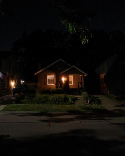 Color photograph looking across a residential street at night at the front of a house, light from a streetlight illuminating the street and front yard and casting dark shadows from overhead tree branches, the front of the house set back in shadow and illuminated by a warmly glowing porch light