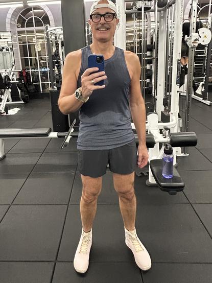 A man (me) is taking a mirror selfie in a gym. He is wearing a gray tank top, dark shorts, and light-colored athletic shoes. A water bottle is visible on a nearby bench. Gym equipment is in the background.